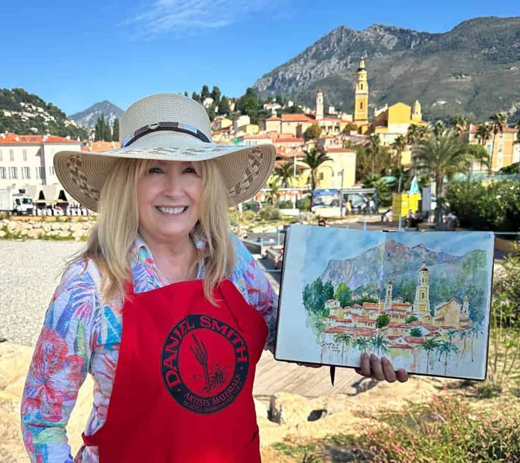 Cindy Briggs in a sun hat and red Daniel Smith apron holding a watercolor sketch of Menton, France, painted on location