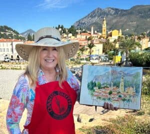 Cindy Briggs in a sun hat and red Daniel Smith apron holding a watercolor sketch of Menton, France, painted on location