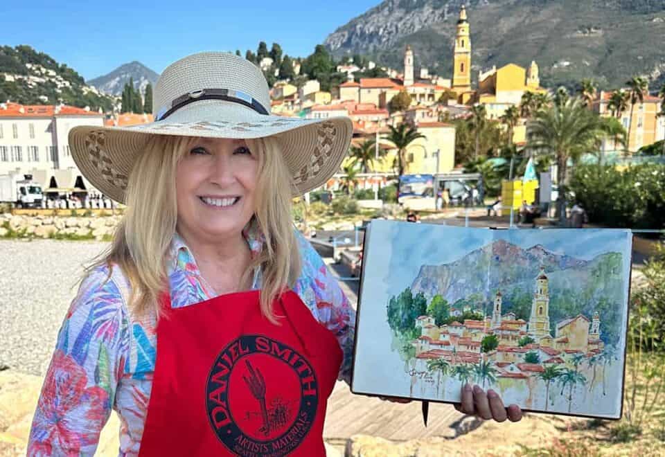 Cindy Briggs in a sun hat and red Daniel Smith apron holding a watercolor sketch of Menton, France, painted on location