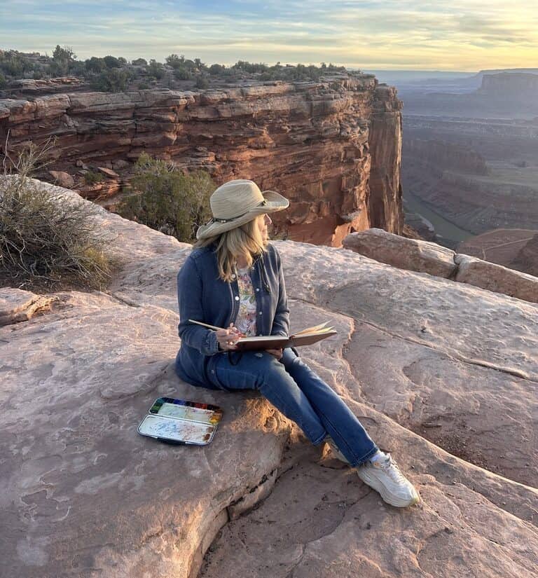 Creativity and Wellness: Why Creativity Feels So Restorative Cindy Briggs Art Watercolor Painting Cindy Briggs sitting on red rock at the edge of a canyon at sunset, sketching in a watercolor journal with her palette beside her
