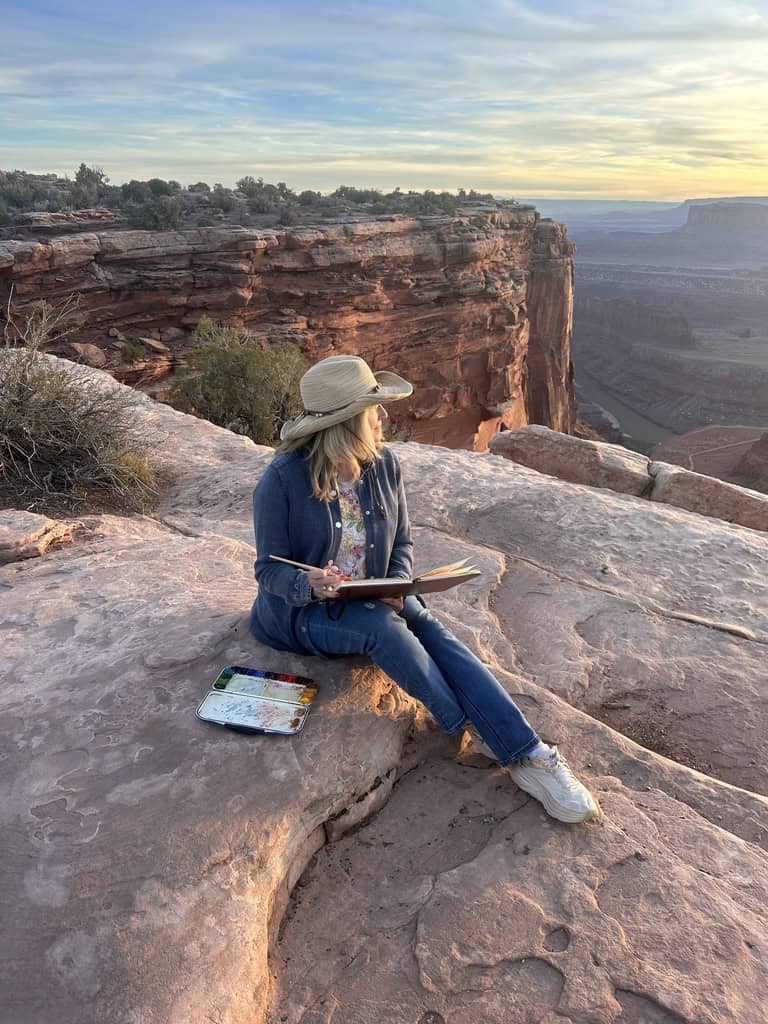 Cindy Briggs sitting on red rock at the edge of a canyon at sunset, sketching in a watercolor journal with her palette beside her