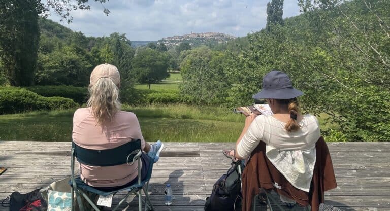 Two women sitting on a wooden dock overlooking a lush green landscape and lake.